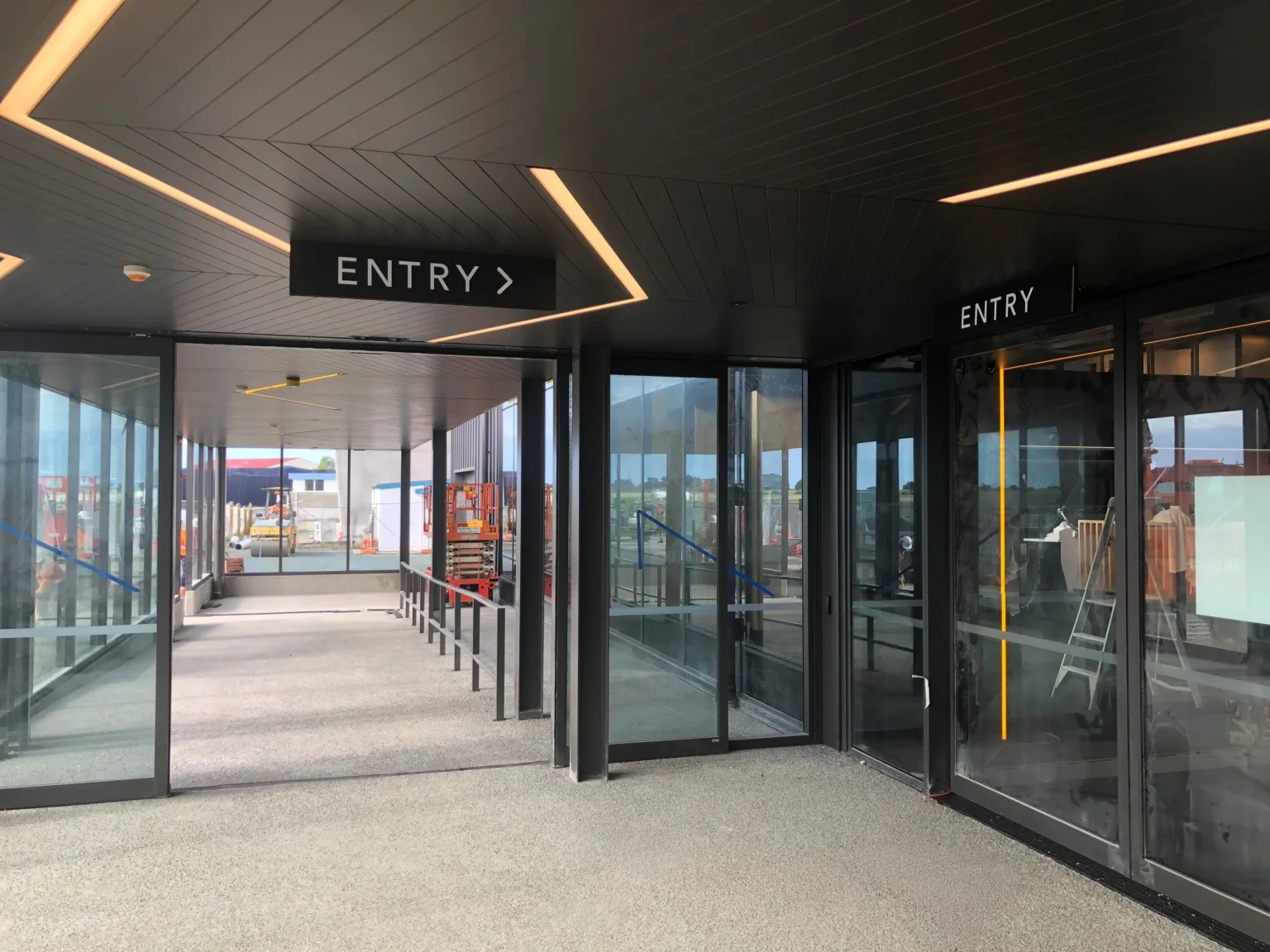 Black under veranda signs with white acrylic lettering reading "entry" and an arrow pointing to the door where there is a second "entry" sign in the same style. Seen fixed to a ceiling with a wooden herringbone patter where some of the slats have been replaced by warm white light strips
